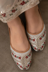 A close-up of a person wearing traditional pointed flat shoes with intricate silver beadwork, red floral embroidery, and green leaf accents, paired with a matching embroidered garment.