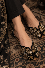 A close-up of a person wearing black flat embellished mule shoes with intricate gold and pearl-like beadwork, standing on a patterned rug next to black embroidered clothing.