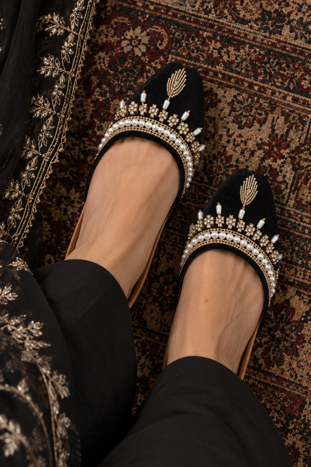 Black velvet flat shoes with gold embroidered motifs and white pearl embellishments, worn on a person's feet over a patterned brown and red rug.